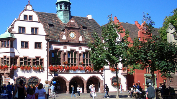 Stadtbild  Ein Platz in einer Kleinstadt mit historischen Gebäuden. Menschen flanieren über dem Platz und sitzen im Schatten unter Bäumen.