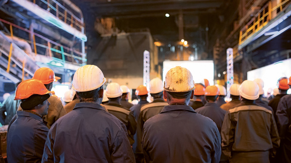 Dargestellt sind Helm tragende Arbeiter von hinten während einer Betriebsversammlung.