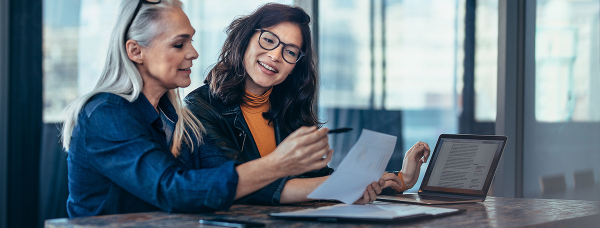 Zwei Geschäftsfrauen im Gespräch an einem Arbeitstisch mit Laptop und Dokumenten.