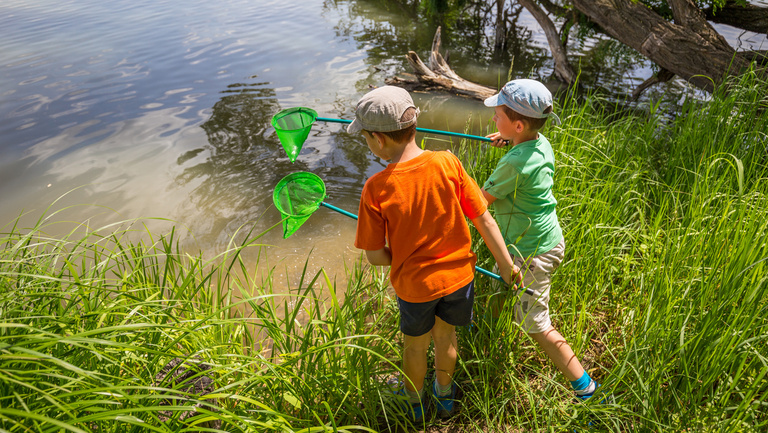 Zwei Jungen stehen am Ufer eines Gewässers und wollen mit einem Köcher im Wasser schöfpen.