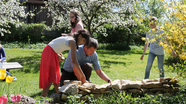 Blüten im Frühling  Ein Mann und eine Frau kümmern sich um eine Trockensteinmauer. Im Hintergrund gehen Kinder durch eine große blühenden Freifläche.