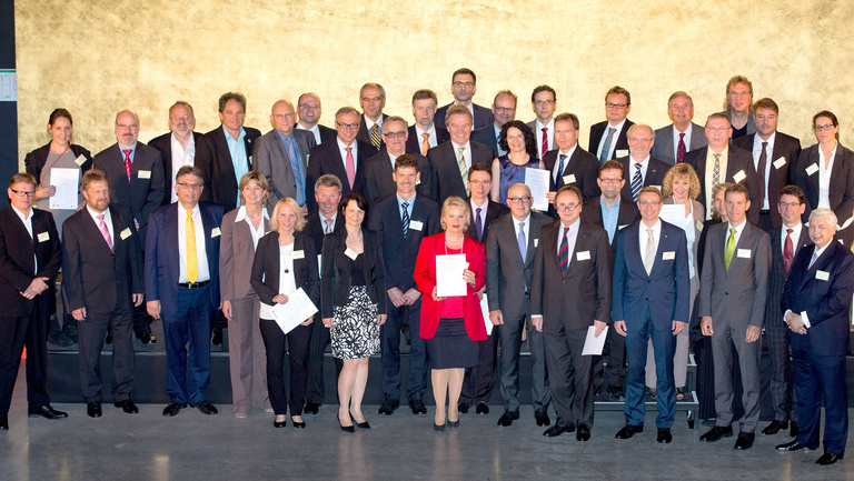 Eine große Gruppe von Unterzeichnerinnen und Unterzeichner der WIn-Charta stehen vor einer goldenen Rückwand zu einem Gruppenfoto zusammen.