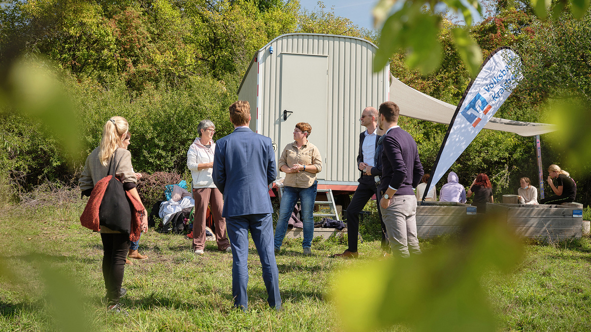 Mehrere Menschen stehen im Kreis auf einer Wiese vor einem Wohnwagen und unterhalten sich. Im Hintergrund sitzen einige Schülerinnen.