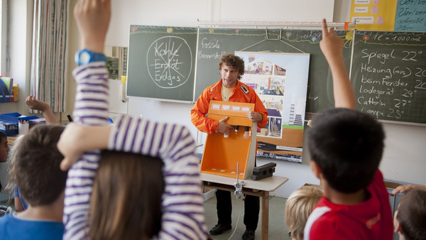 Energie-Detektiv EDe - ein außerschuliches Angebot Energie-Detektiv EDe steht mit seiner orangefarbenen Jacke von der Tafel in einem Klassenzimmer und erklärt den Schulkindern einen Lampenkasten, mit dem die Kinder verschiedene Beleuchtungsmittel auf Stromverbrauch und Lichtausbeute untersuchen können. Im Vordergrund sient man ein paar Kinder, die sich eifrig zu Wort melden wollen.