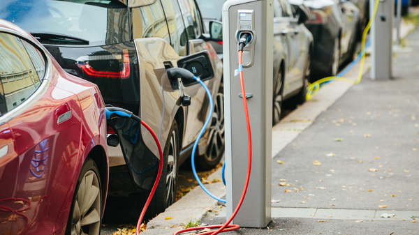Das Bild zeigt mehrere Elektro-Autos am Straßenrand, die an einer Elektroladesäule angeschlossen sind.
