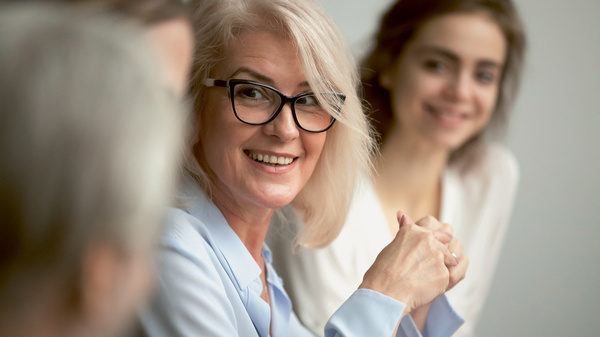 Dargestellt ist eine Besprechung, im Vordergrund eine etwas ältere Frau, die jemandem zuhört. Im Hintergrund eine jüngere Frau.