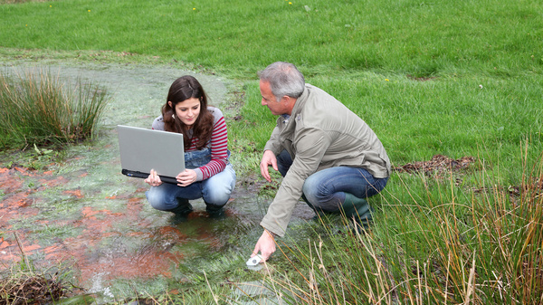 Wissenstransfer Eine Jugendliche und ein älterer Mann entnehmen auf einer überschwemmten Wiese eine Wasserprobe. Die junge Frau hat einen Laptop in den Händen.