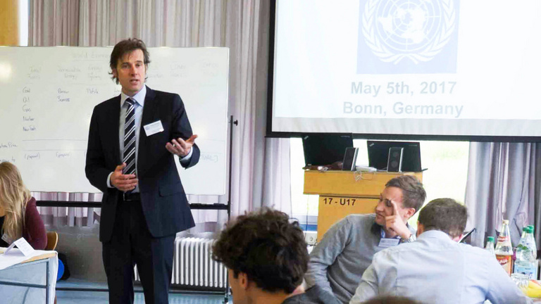 Mehrere junge Erwachsene an zwei Tischen, die dem vortragenden Mann in der Mitte des Raums zuhören