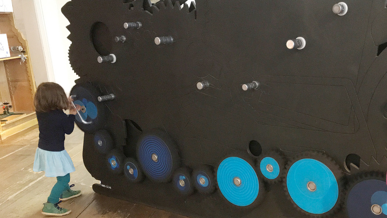 Ein Mädchen steht an einer Wand und dreht eine selbst zusammengestellte Zahnradkette.