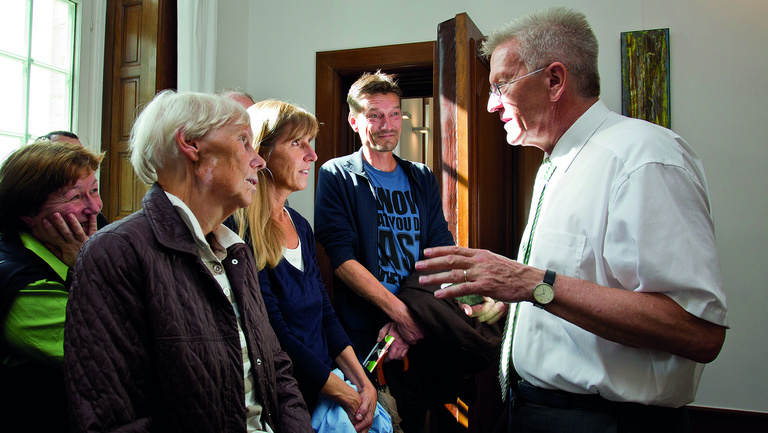 Ministerpräsident Winfried Kretschmann erklärt einer Gruppe von sieben Leuten etwas in einem hellen Raum. 