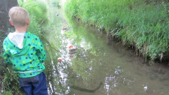 Ein Junge steht am Ufer eines schmalen Flusses, auf dem mehrere kleine selbstgebaute Schiffchen schwimmen.