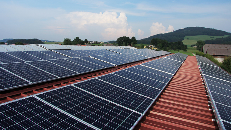 Vier lange Reihen mit Solarzelle bedecken das Dach einer großen Halle, die in einer dörflichen Umgebung steht.