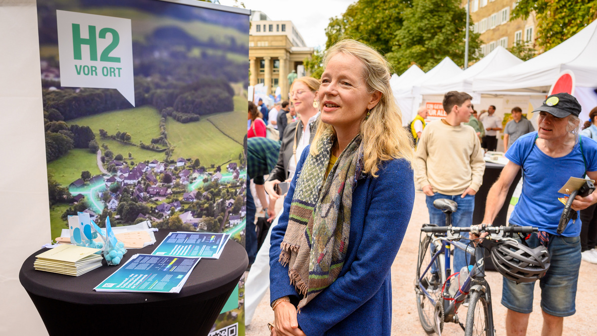 Thekla Walker, Ministerin für Umwelt, Klima und Energiewirtschaft, steht an einem Stehtisch vor einem Plakat mit dem Logo "H2 vor Ort" und der Abbildung eines ländlichen Dorfes vor oben.