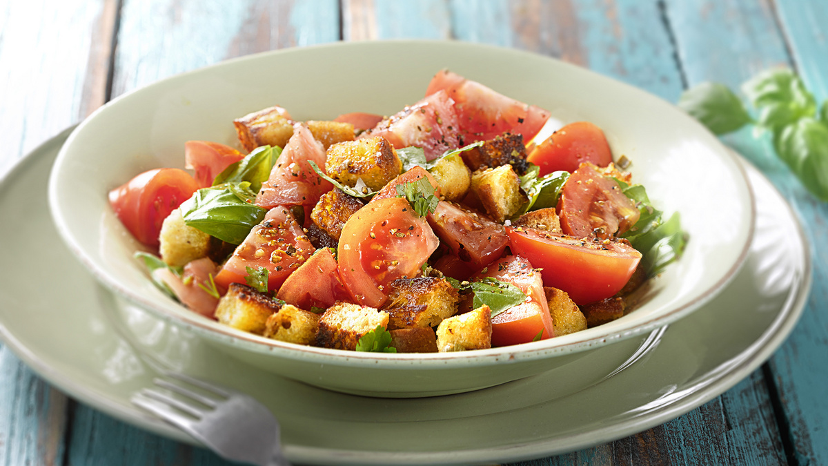 Auf einem blaugestrichenen Holztisch steht ein Teller mit Brotsalat aus Brotresten, Tomaten und Kräutern.