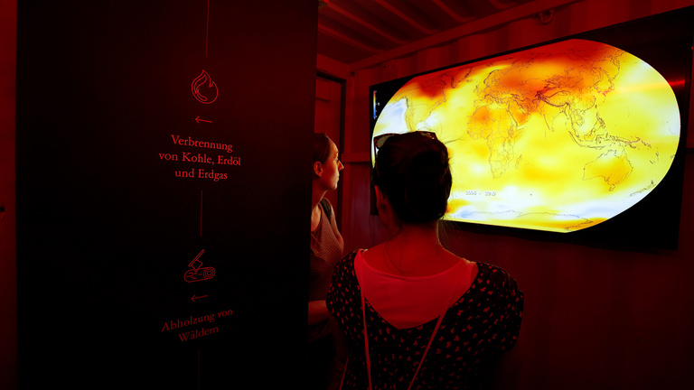 Eine Ausstellung zum Thema präsentiert auf einem Monitor die Entwicklung der Temperaturen auf der Erde von 2006 bis 2010. Zwei Besucherinnen betrachten den Monitor, seitlich befindet sich eine Informationswand.