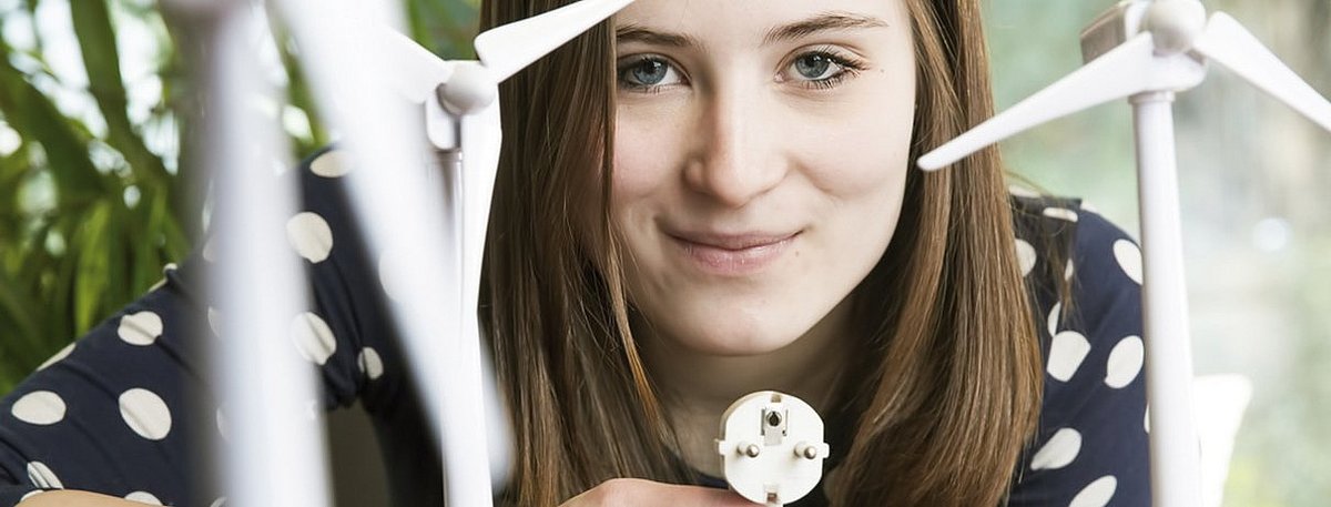 Eine junge Frau sitzt hinter einem Tisch. Vor ihr stehen drei Modell-Windräder, in der Hand hält sie eine Schutzkontaktstecker.