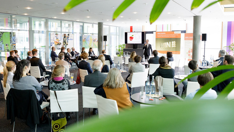 Interessierte und Unternehmen sitzen während WIN Forum an Tischen und schenken einem Vortrag ihre Aufmerksamkeit.