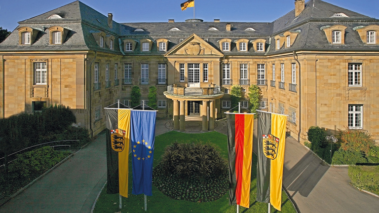 Die Villa Reitzenstein ist ein antikes, prachtvolles Gebäude mit einer Steinfassade und grauem Dach. Vor ihr sind vier verschiedene Fahnen gehisst. Unter anderem die europäische, die deutsche und die baden-württembergische Flagge.