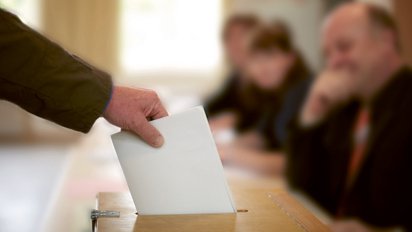 Dargestellt ist eine Wahlurne, in die eine Person seine Wahlunterlagen hineinwirft.