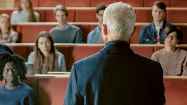 Dargestellt ist ein Hörsaal. Eine Lehrperson spricht zu Studenten und Studentinnen.