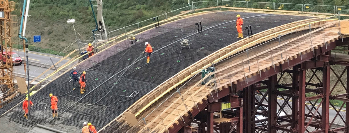 Baustelle einer Brücke mit mehreren Arbeitern in orangefarbener Schutzkleidung, Stahlgerüste und ein Betonpumpenarm sichtbar.