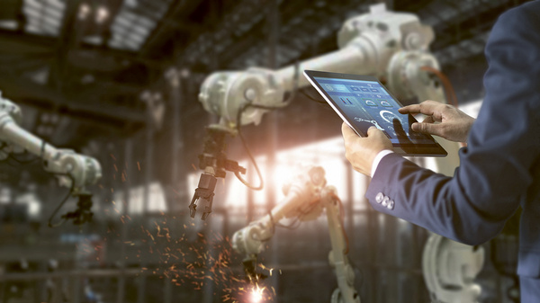 Dargestellt ist ein Fertigungsroboter in einer Fabrik. Eine Person steuert die Maschine mithilfe eines Tablets.