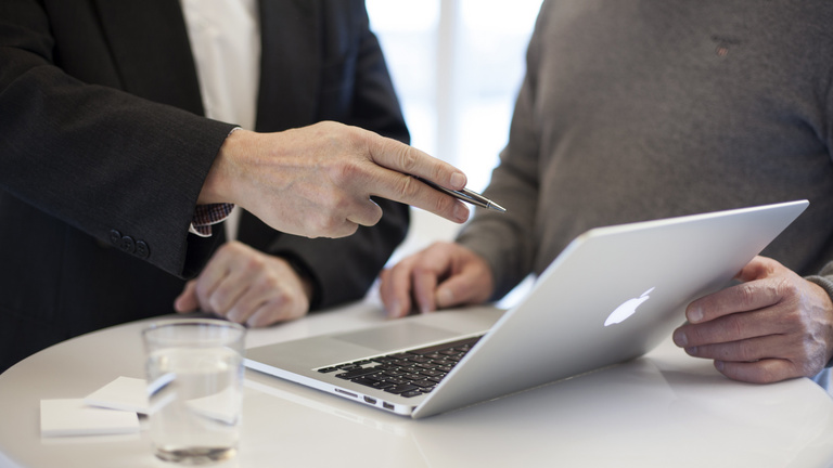 Beratung Im Mittelpunkt des Bildes befindet sich ein Laptop, weiter sind von zwei Personen der Rumpf zu sehen. Beide befassen sich mit dem Inhalt auf dem Bildschirm.