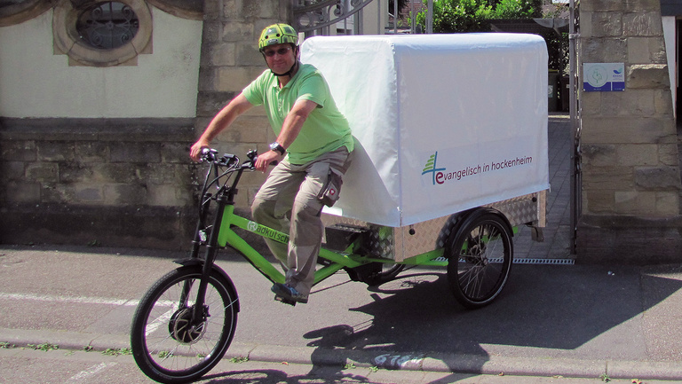 Lastenfahrrad der Evangelische Kirchengemeinde Hockenheim Ein Mann fährt mit einem Lastenfahrad, das hinten seine Ladefläche hat, aus einer Hofeinfahrt heraus.
