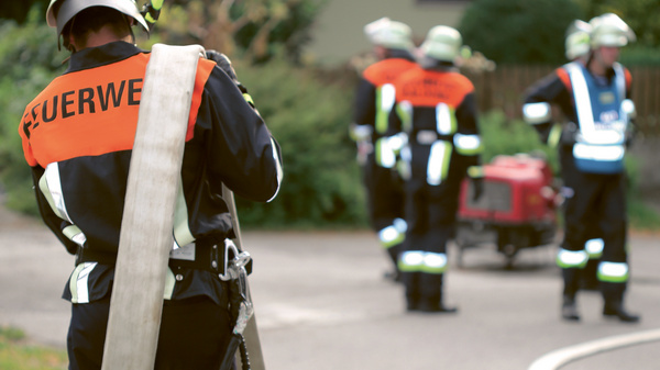 Dargestellt sind Feuerwehrleute im Einsatz. Einer der Feuerwehrmänner trägt einen Schlauch über der Schulter.