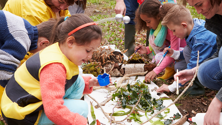 Kinder und mehrere Erwachsene breiten Blätter und anderes Naturmaterial auf einer weißen Fläche aus. Ein Mädchen pinselt.