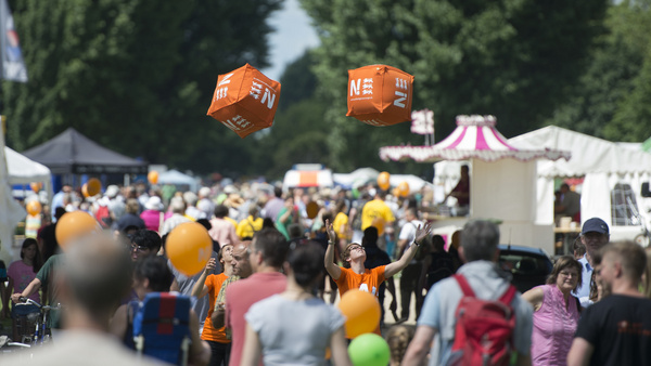 N!-Tour 2013 Vielen Menschen tummeln sich bei einer Veranstaltung der Nachhaltigkeitsstrategie. Zwei orangefarbene Würfel fliegen schweben über den Besuchern.