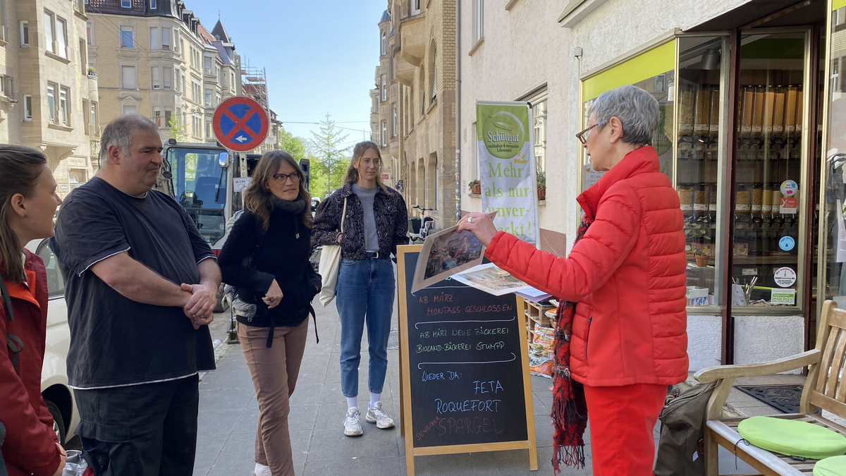Methodenwerkstatt Stadtführung 2024 Menschen hören einer Referentin zu, sie stehen vor einem Laden im Rahmen einer Stadtführung