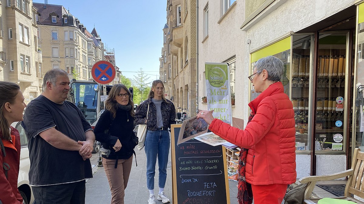 Methodenwerkstatt Stadtführung 2024 Menschen hören einer Referentin zu, sie stehen vor einem Laden im Rahmen einer Stadtführung