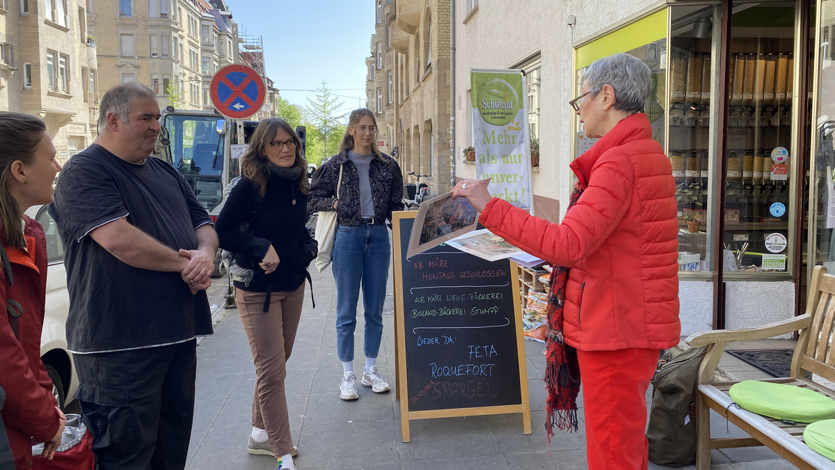 Methodenwerkstatt Stadtführung 2024 Menschen hören einer Referentin zu, sie stehen vor einem Laden im Rahmen einer Stadtführung